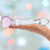 A hand holds a clear glass object with a pink, heart-shaped handle, set against a blurred background of colorful lights. The Gläs 8" Sweetheart Glass Dildo reflects light beautifully and has a smooth, glossy surface.